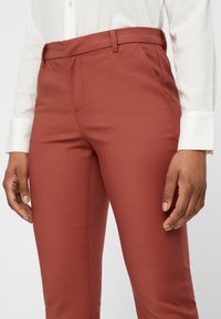 Rust-colored tailored pants with a smooth, structured fabric and sleek front pockets. Belt loops visible at the waistline.