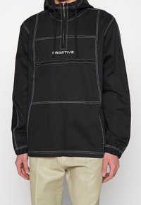 Polo noir à capuche avec coutures contrastantes blanches et logo "Primitive", poche poitrine zippée, porté avec un pantalon beige clair.