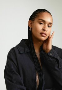 Svart denimjacka med krage, bärs över en beige topp. Personen har långt flätat hår och en silverfärgad hjärtformad örhänge.
