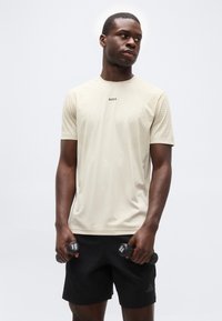 Άνδρας με μπεζ μπλουζάκι BOSS και μαύρο σορτς κρατά δύο βαράκια 1 κιλού, κοιτάζοντας πλάγια με φόντο μονόχρωμο.