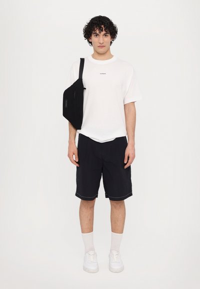 Weißes Baumwoll-T-Shirt mit kleinem Branding, schwarze Shorts, weiße Sneaker und eine schwarze Umhängetasche. Die Tasche hat einen Reißverschluss und Logo-Details.
