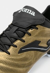 Μαύρα και χρυσά ποδοσφαιρικά παπούτσια με υφασματική επιφάνεια, διαθέτοντας μαύρη επικάλυψη κορδόνων, επενδεδυμένη γλώσσα και λευκό λογότυπο.