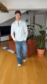 Camisa blanca de manga larga con cuello y un detalle rojo, combinada con jeans azules sueltos y zapatos blancos, en un ambiente hogareño con plantas.