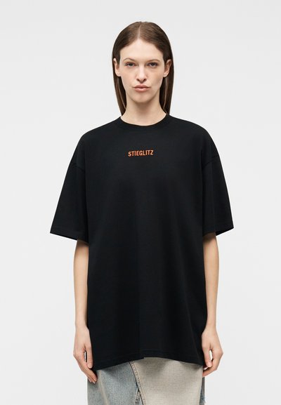 Γυναίκα με ίσια καστανά μαλλιά που φοράει άνετο μαύρο Τ-shirt με πορτοκαλί κείμενο "STIEGLITZ" στο κέντρο του στήθους και φούστα ανοιχτού χρώματος.