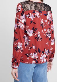 Röd blus med långa ärmar, prydd med en svart spetsbalk, strukturerad tyg och ett mönster av rosa och marinblå blommor.