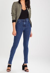 Chaqueta bomber de color oliva sobre top negro, jeans skinny de tiro alto en azul con dobladillo deshilachado, y sandalias negras de tacón bloque.