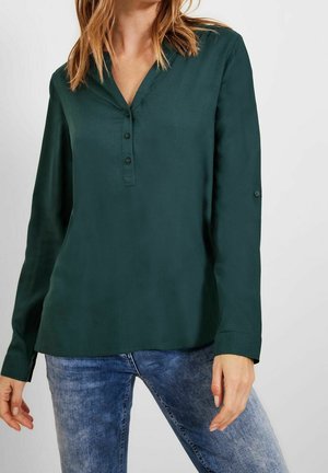 Kobieta ubrana w ciemnozieloną bluzkę z długim rękawem i dekoltem w serek zapinaną na guziki, w połączeniu z wyblakłymi niebieskimi dżinsami na jednolitym tle.