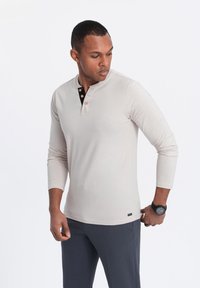 Beige Langarm-Henley-Shirt mit einer schwarzknöpfigen Knopfleiste; glatte Textur; getragen mit dunkelgrauen Hosen; Armbanduhr sichtbar.