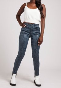Jeans de cintura alta, con estampado de serpiente azul y un diseño ajustado, combinados con una camiseta cropped blanca y botas negras hasta el tobillo.