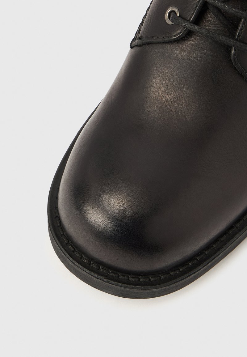 Chaussure en cuir noir avec une texture lisse et brillante, bout arrondi et bord festonné. Dispose de lacets fins et d'œillets métalliques sur le col.