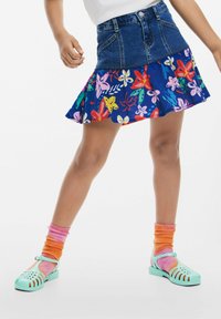 Gonna in denim e floreale con orlo a balze, caratterizzata da colori vivaci e fantasie. Indossata con scarpe verde menta e Calzini multicolori.