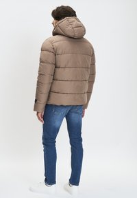 Chaqueta de plumón marrón con costuras horizontales, capucha y un corte relajado, combinada con vaqueros azules y zapatillas blancas. Vista trasera.