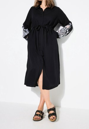 Donna che indossa un vestito midi nero con dettagli in pizzo bianco sulle maniche, vita annodata e sandali neri con doppia fibbia borchiata, in piedi contro un muro bianco.