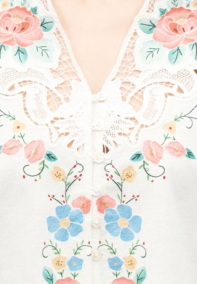 Λευκή μπλούζα με πολύχρωμα floral μοτίβα και λεπτομέρειες δαντέλας γύρω από το ντεκολτέ, με μικρά λευκά κουμπιά στο κέντρο.