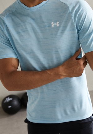 Mand iført lyseblå Under Armour t-shirt med korte ærmer, trækker ærmet med den ene hånd, fitnessudstyr sløret i baggrunden.