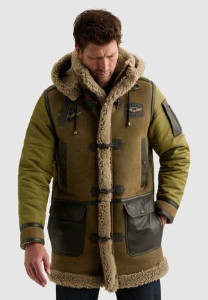 Homme portant un parka en shearling marron et vert avec de grandes poches avant et une capuche, regardant vers le bas avec les mains près des poches.