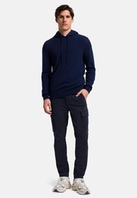 Felpa blu navy con cappuccio regolabile, abbinata a pantaloni cargo scuri con tasche laterali. Modello con scarpe sportive chiare.
