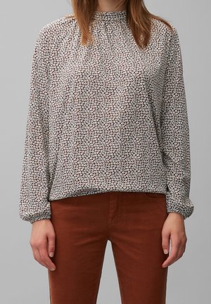 Kvinde iført en langærmet bluse med blomsterprint kombineret med rustfarvede bukser, stående mod en ensfarvet grå baggrund.