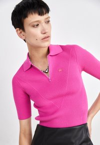 Enganpasst, geripptes, pinkes Polo-Shirt mit einem Reißverschluss vorn, kurzen Ärmeln und einem kleinen, geprägten Logo. Kombiniert mit einem schwarzen Lederrock.