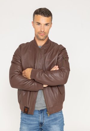 Man met kort haar en lichte baard, gekleed in een bruin leren bomberjack over een grijs shirt en blauwe spijkerbroek, met gekruiste armen, neutrale achtergrond.