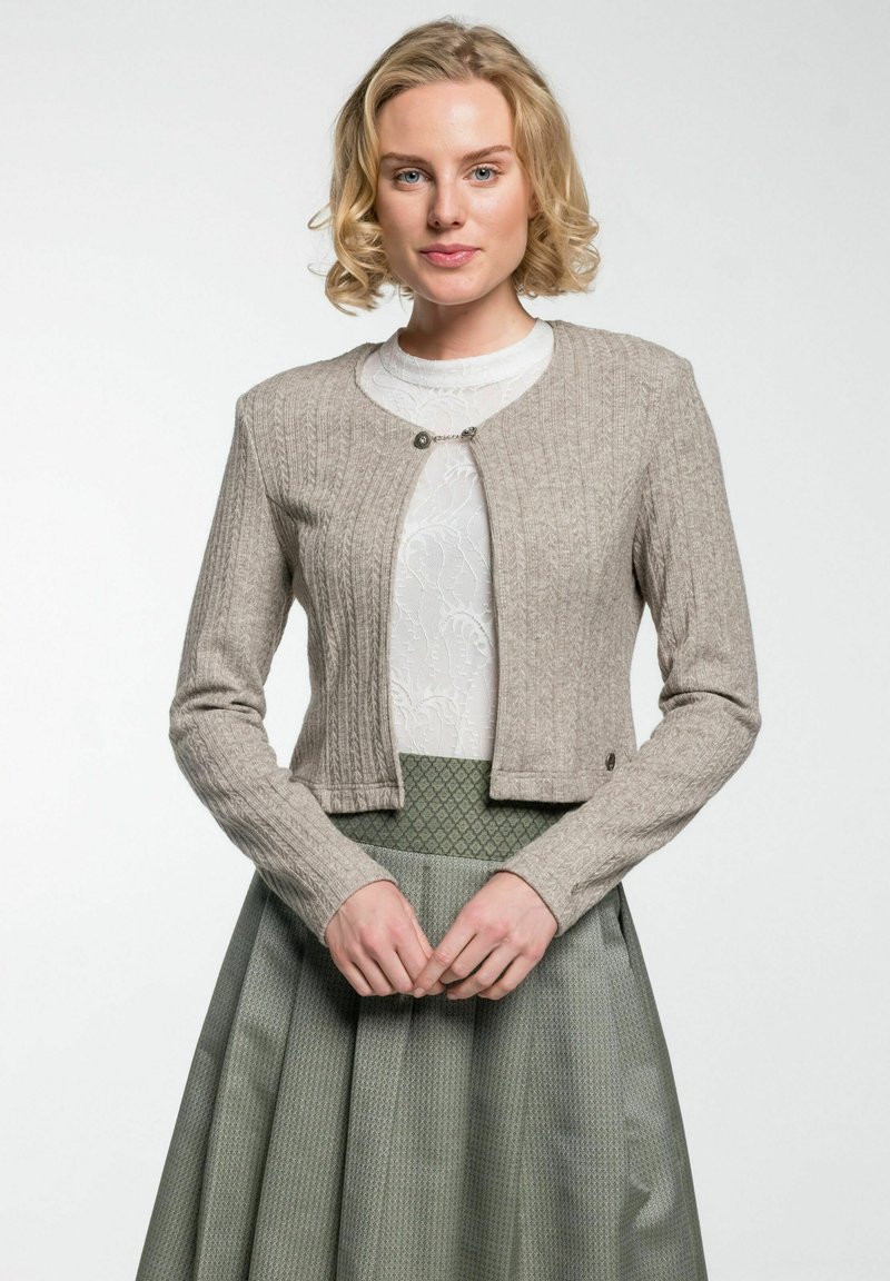 Beige gestrickte Strickjacke mit Rippmuster, cropped Länge, Rundhalsausschnitt und silberner Knopfverschluss, darüber getragen über einem weißen Spitzenoberteil und einem grünen Rock.