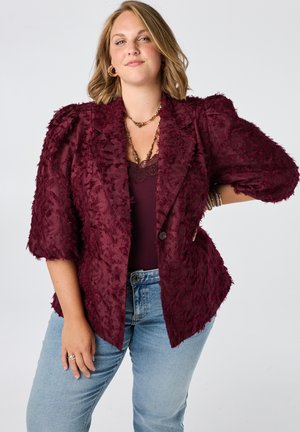 Vrouw met schouderlang blond haar draagt een gestructureerd bordeauxblauw blazer, bijpassende top, lichtblauwe spijkerbroek en gouden sieraden tegen een effen achtergrond.