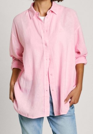 Henkilö pukeutunut väljään, vaaleanpunaiseen napitettuun paitaan, jonka hihat on kääritty, valkoisen paidan päällä, yhdistettynä vaaleansinisiin farkkuihin, kädet taskuissa.