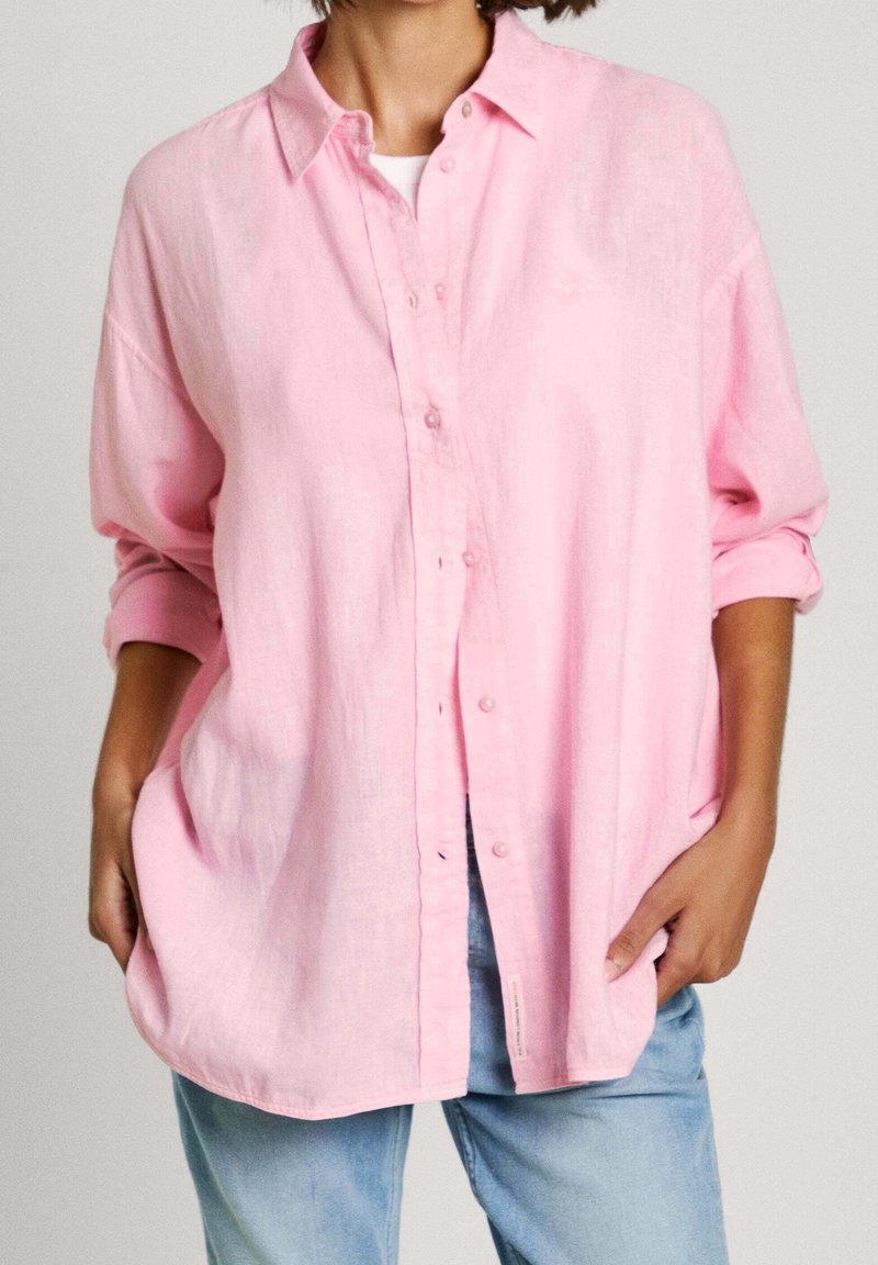 Personne portant une chemise ample rose clair à boutons avec manches retroussées sur un haut blanc, associée à un jean bleu clair, mains dans les poches.