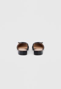 Ζευγάρι μαύρα δερμάτινα σανδάλια με επίπεδη σόλα και λουριά αστραγάλου, ορατά από πίσω, σε απλό γκρι φόντο.