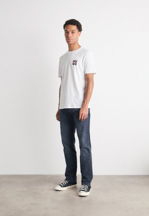 Camiseta blanca de algodón con el logo "HUGO" en rojo y negro, combinada con jeans de mezclilla azul oscuro y zapatillas negras con suelas blancas.