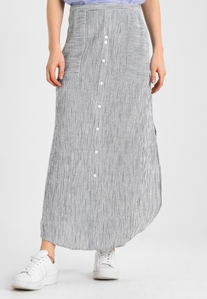 Femme portant une longue jupe boutonnée à rayures verticales noires et blanches avec deux poches avant et des baskets blanches.