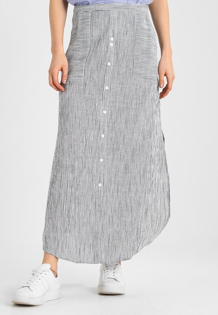 Femme portant une longue jupe boutonnée à rayures verticales noires et blanches avec deux poches avant et des baskets blanches.