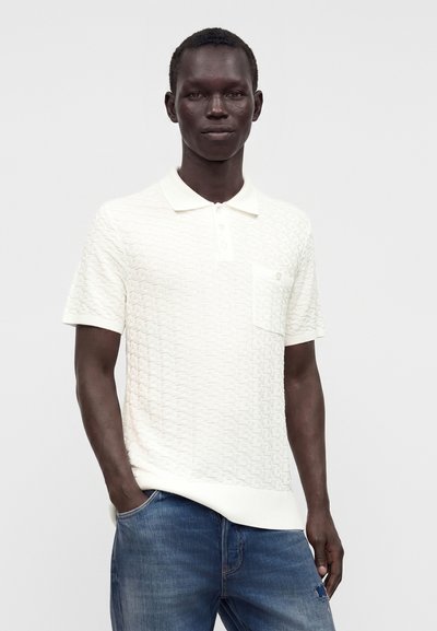 Homme debout portant un polo blanc texturé à manches courtes et un jean bleu avec une main dans la poche, devant un fond uni.