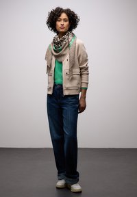 Femme aux cheveux bouclés portant une veste beige, un pull vert, une écharpe à imprimé animal, un jean bleu et des baskets blanches, debout devant un fond uni.