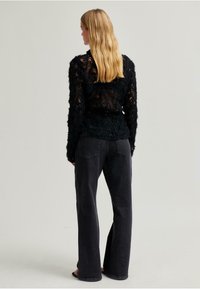 Černý pletený top s texturou a dlouhými rukávy, kombinovaný s tmavými džíny s vysokým pasem. Vlasy jsou vlnité a sahají přes ramena.