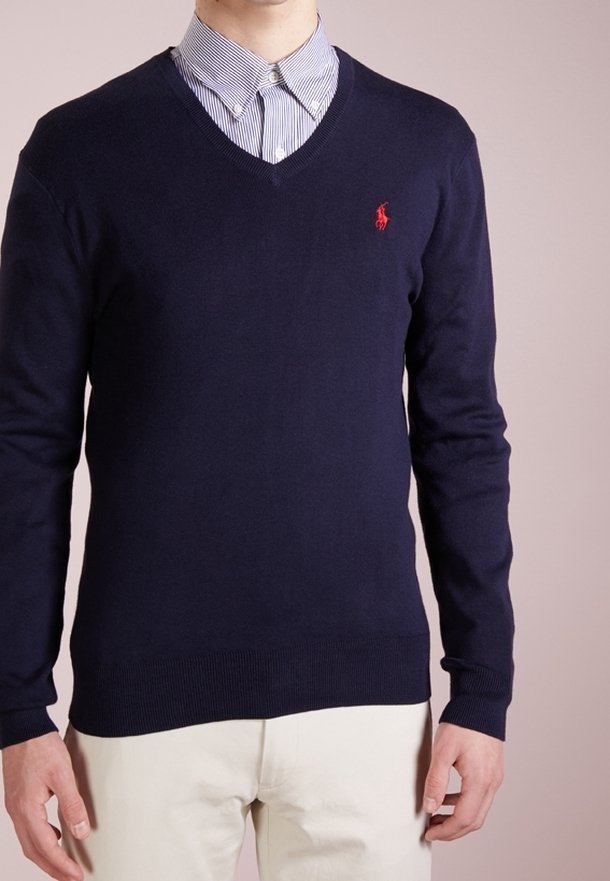 Navyblauer V-Ausschnitt-Pullover aus weichem Material, mit langen Ärmeln und einem kleinen roten Logo auf der Brust, getragen über einem gestreiften Kragenhemd.