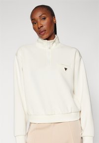 Crèmekleurige half-zip trui met elastische manchetten en boord, met een klein driehoekig logo op de borstzak. Zachte stoftextuur.