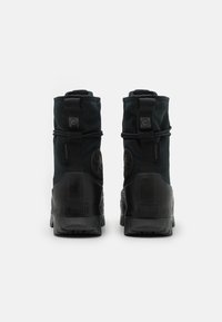 Schwarze wasserdichte Knöchelstiefel mit einer langlebigen Gummisohle, strukturiertem Obermaterial und Markenemblem auf der Rückseite. Mit verstellbaren Zugriemen.