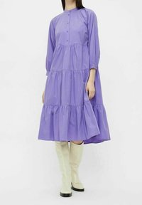Vestido de algodón morado, de capas, con mangas largas y cierre de botones en la parte delantera. El vestido tiene una silueta fluida y se combina con botas blancas hasta la rodilla.