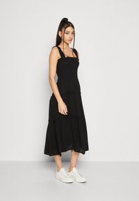 Robe maxi noire à volants avec des bretelles ajustables et un corsage ajusté, arborant une silhouette fluide. Portée avec des baskets blanches pour un contraste.
