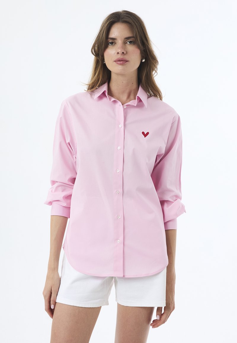 Chemise boutonnée rose clair à manches longues, ornée d'une petite broderie en forme de cœur rouge sur la poitrine. Texture du tissu en coton lisse.