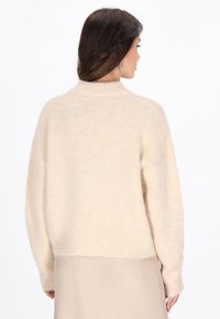 Pulover tricotat de culoare crem, cu guler înalt, croială relaxată și material texturat. Prezintă ornamente cu perle pe mâneci. Moale și ușor.
