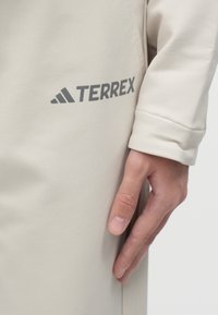 Beige TERREX dzseki texturált anyagból, hajtott ujjú, szürke nyomott logóval az oldalán. Kéz az anyagon.