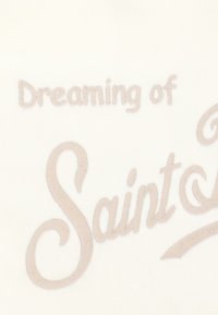 Il testo ricamato recita "Dreaming of Saint L," su uno sfondo di tessuto liscio color crema. La cucitura è in beige chiaro.