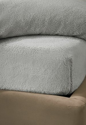 Couverture en polaire douce et gris clair, pliée et placée dans le coin d’un canapé ou fauteuil beige rembourré.