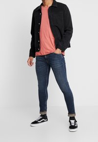 Svart denimjacka över en korallfärgad t-shirt, kombinerad med slim fit blå jeans och svarta sneakers. Noterbara detaljer inkluderar upprullade ärmar och vita skodetaljer.