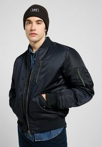 Giacca bomber nera con polsini e bordo a costine, presenta numerose tasche con zip e una consistenza liscia e lucida. Indossata sopra una camicia di denim.