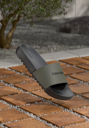 Infradito Calvin Klein neri e verde oliva che galleggiano sopra un pavimento in piastrelle di legno con piccoli ciottoli in un ambiente all'aperto.