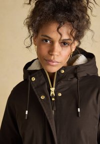 Parka marron avec capuche, fermeture éclair dorée et boutons-pression. Comprend une doublure douce et texturée ainsi que des cordons de serrage au niveau du col et de l'ourlet.