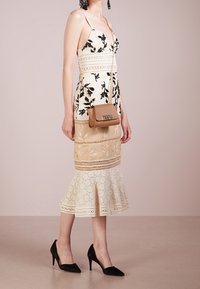 Abito floreale ricamato bianco e beige con dettagli in pizzo e orlo svasato, abbinato a tacchi a spillo neri e una borsa marrone.
